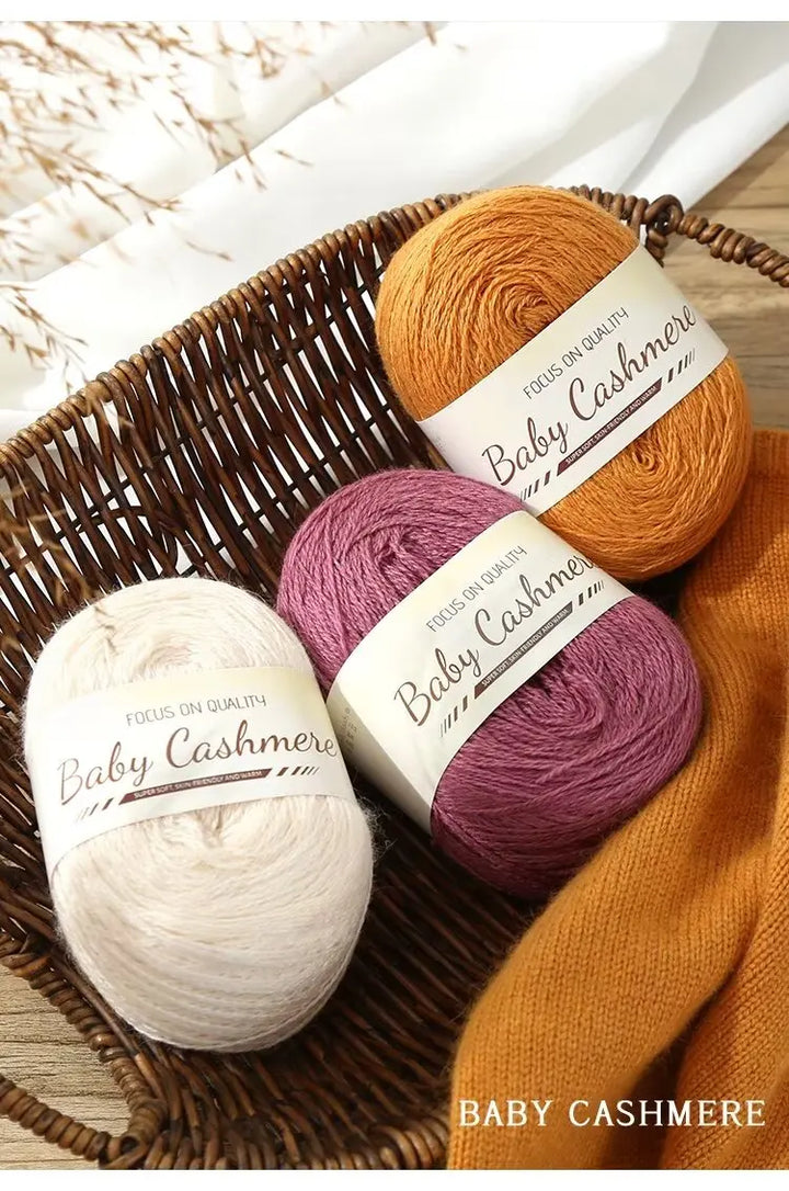 Three balls of baby cashmere yarn in orange, purple, and white on a woven basket.
