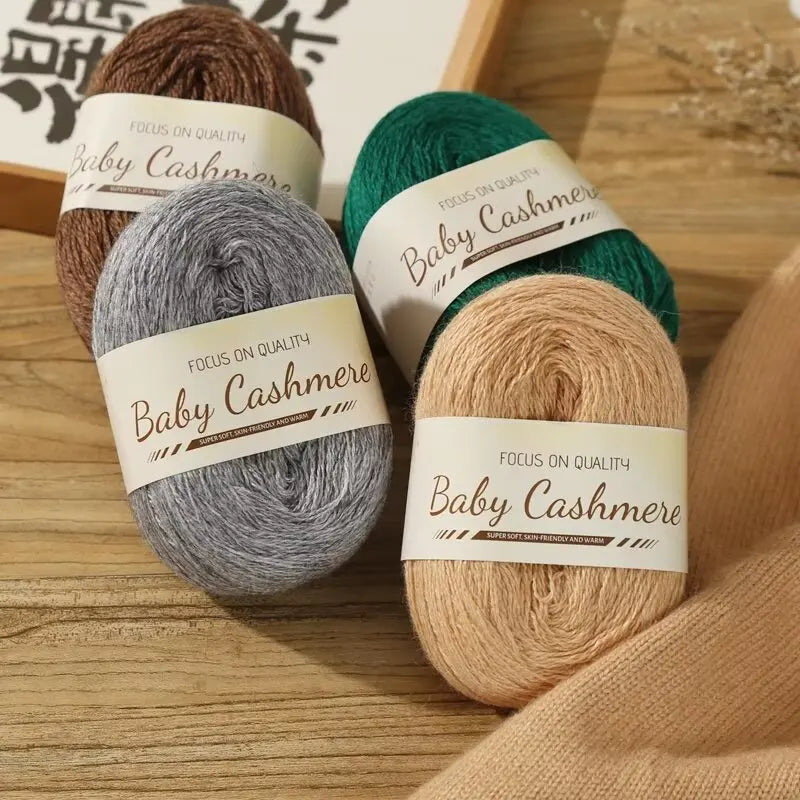 Four balls of baby cashmere yarn in brown, green, gray, and beige on a wooden surface with 'Baby Cashmere' labels.